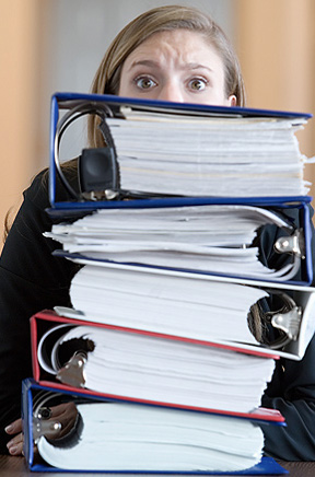 A worried looking woman sitting behind a stack of files with nothing but her eyes looking out over the top of the files.
